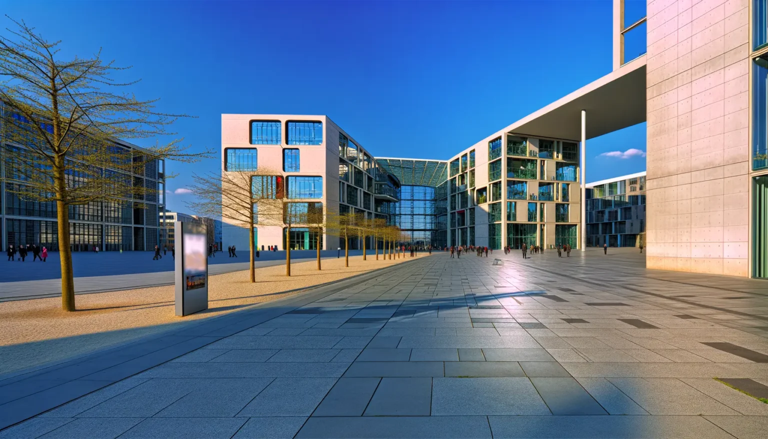 Moderner Platz mit futuristischen Gebäuden, großen Fensterfronten und einer geräumigen, gepflasterten Fläche, Menschen gehen im Abstand unter klarem blauem Himmel.