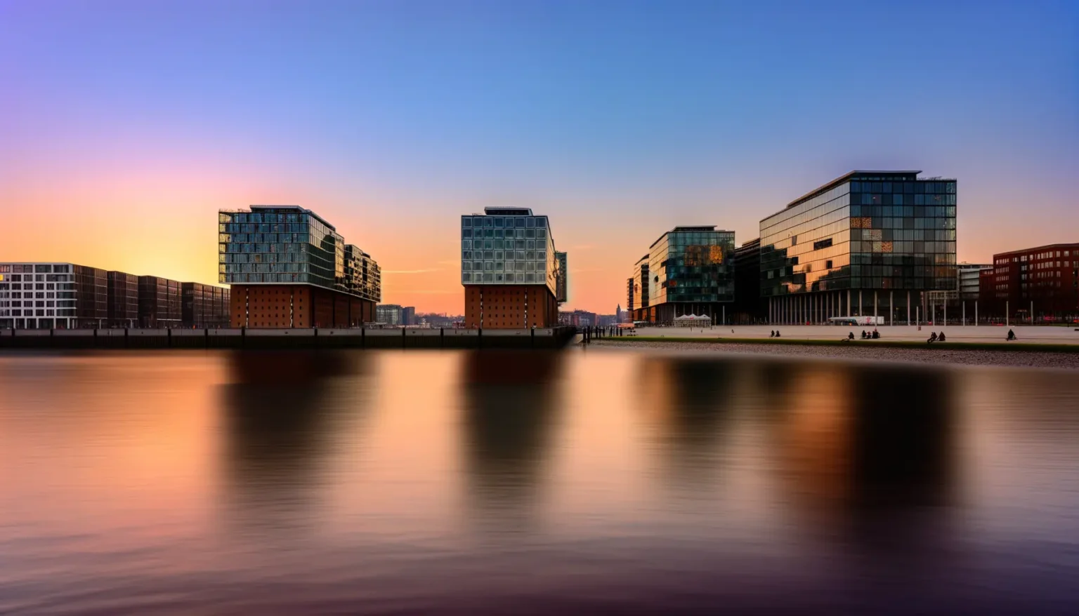Moderne Gebäude mit Glasfassaden entlang eines ruhigen Gewässers bei Sonnenuntergang mit einem farbenfrohen Himmel von Orange bis Blau. Die Architektur spiegelt sich weich im Wasser wider, und einige Menschen entspannen sich am Ufer.