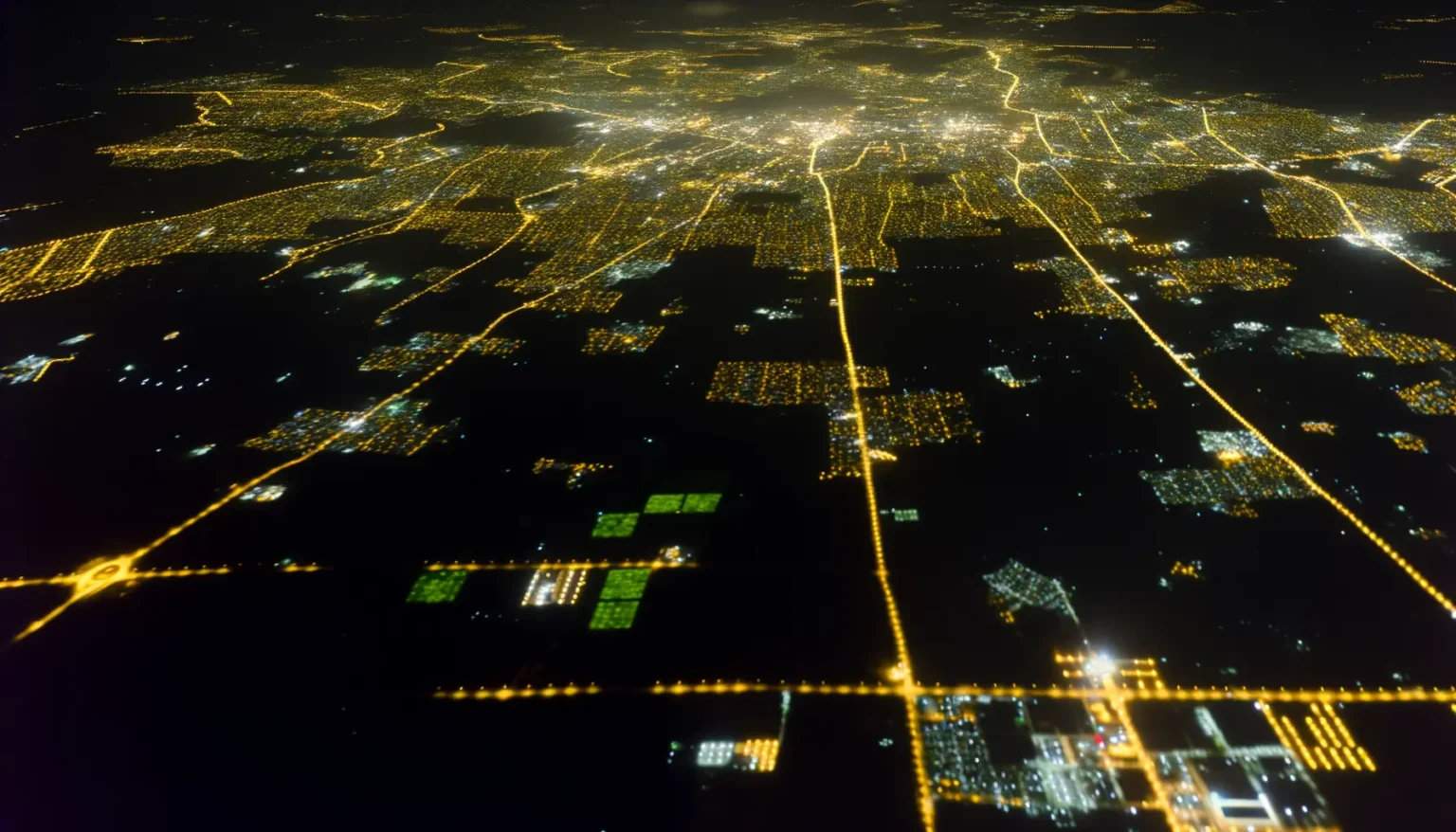 Luftaufnahme einer Stadt bei Nacht, mit zahlreichen goldenen Lichtern, die Straßen und dicht bebaute Gebiete markieren, umgeben von dunklen Flächen mit vereinzelten Lichtern, die weniger dicht besiedelte oder unbebaute Gebiete andeuten. Die Lichter bilden ein komplexes Muster, das auf eine große urbane Ausdehnung hinweist.