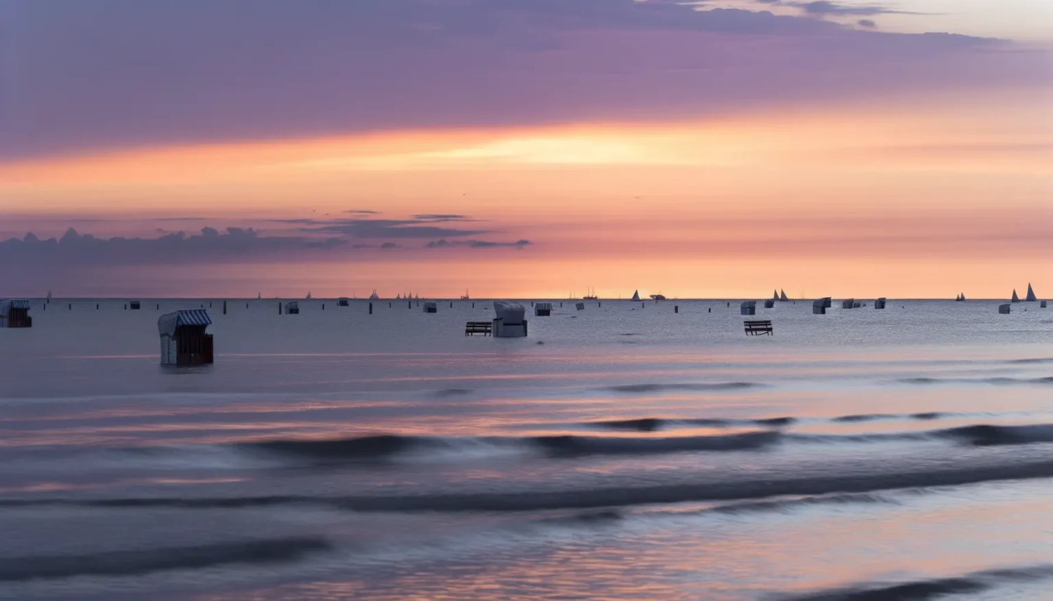 Ein friedvoller Sonnenuntergang am Meer mit einem pastellfarbenen Himmel in Rosa- und Orangetönen. Im ruhigen Wasser sind zahlreiche Segelboote in der Ferne zu erkennen und im Vordergrund einige überflutete Strandhütten.